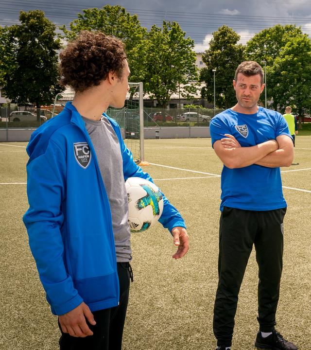 "Notruf Hafenkante - Fußball-Mütter": Noah Kruse (Jörg Rohde), Dominik Vogt (Riccardo Campione) und Lennard Kröger (Luke Matt Röntgen) stehen zusammen auf einem Fußballplatz. Dominik hat einen Fußball unter seinem linken Arm klemmen. Er blickt zu Noah, der links neben ihm steht. Noah hat die Arme vor dem Körper verschränkt und sieht Dominik an. Lennard steht Dominik gegenüber. Er hat die Arme in die Hüften gestemmt und sieht zu Noah.