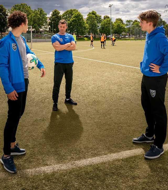 "Notruf Hafenkante - Fußball-Mütter": Noah Kruse (Jörg Rohde), Dominik Vogt (Riccardo Campione) und Lennard Kröger (Luke Matt Röntgen) stehen zusammen auf einem Fußballplatz. Dominik hat einen Fußball unter seinem linken Arm klemmen. Er blickt zu Noah, der links neben ihm steht. Noah hat die Arme vor dem Körper verschränkt und sieht Dominik an. Lennard steht Dominik gegenüber. Er hat die Arme in die Hüften gestemmt und sieht zu Noah.
