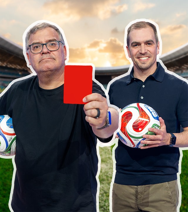 Das Bild zeigt Moderator Elton und den ehemaligen Profifußballer Philipp Lahm in einem Fußballstadion. Elton steht im Vordergrund, hält einen Fußball unter dem Arm und streckt eine rote Karte in die Kamera, während Lahm daneben steht, einen Ball in den Händen hält und freundlich lächelt.