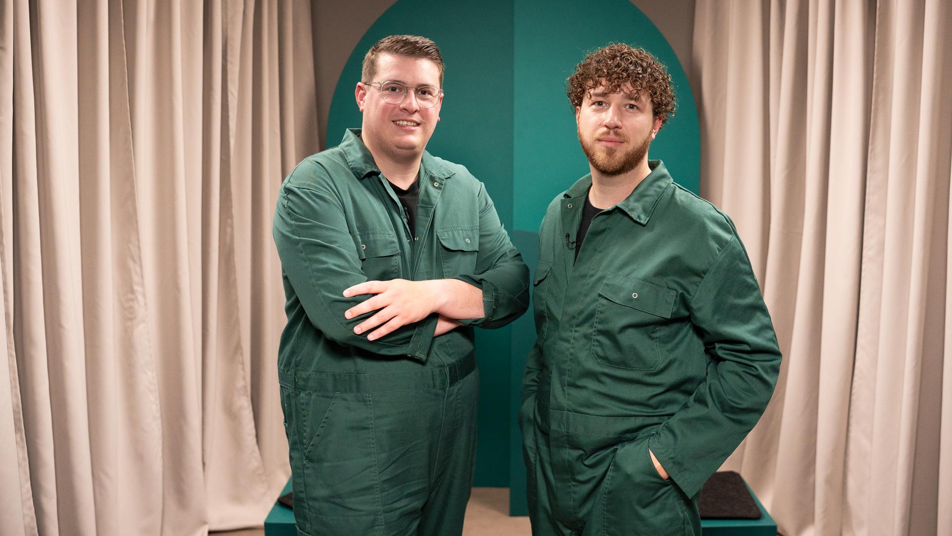 Die beiden Gäste der Sendung tragen grüne Overalls und blicken in die Kamera.