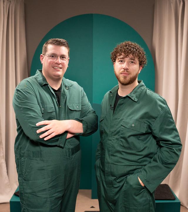 Die beiden Gäste der Sendung tragen grüne Overalls und blicken in die Kamera.