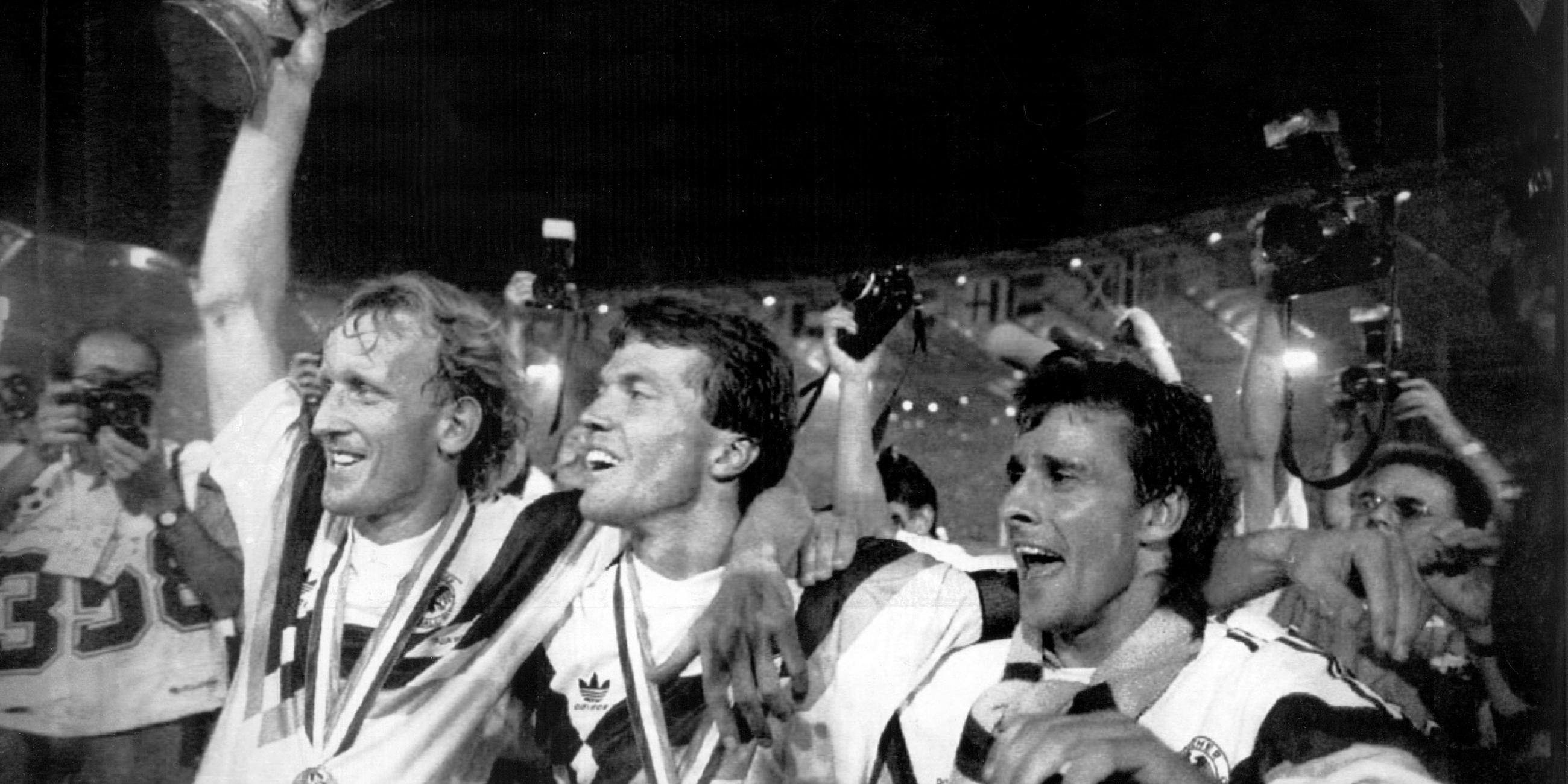 08.07.1990, Italien, Rom: Die damaligen deutschen Nationalspieler Andreas Brehme (l-r), mit dem WM-Pokal in der Hand, Lothar Matthäus und Pierre Littbarski bejubeln den Erfolg im Olympiastadion in Rom.
