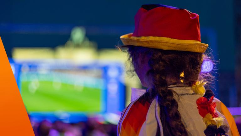Im Vordergrund steht eine Person mit Deutschlandhut und Fanbekleidung, die auf eine unscharfe Großbildleinwand mit einem Fußballspiel blickt. Die Szene spielt in einer stimmungsvollen Umgebung mit vielen Lichtern, typisch für Public Viewing. Das Motiv symbolisiert die wirtschaftlichen Effekte der WM durch gesteigerte Nachfrage nach Fanartikeln, Technik und Gastronomie.