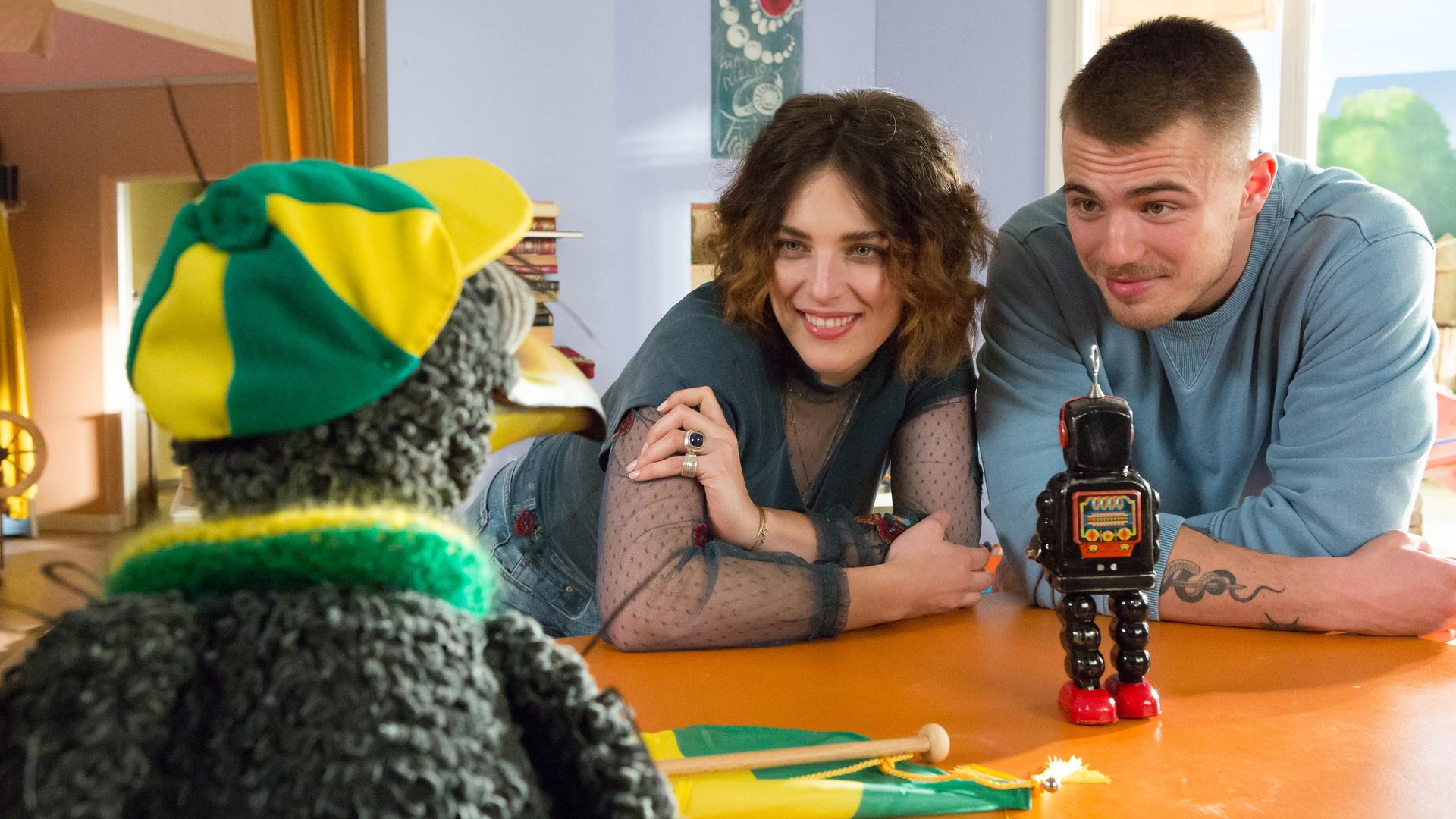 Rudi trägt eine gelb-grüne Kappe und einen gelb-grünen Schal, vor ihm auf dem Tresen legt ein gelb-grüner Wimpel. Er schaut zu Siebenstein und Sommi, vor denen ein Spielzeugroboter steht und die Rudi freundlich anblicken.