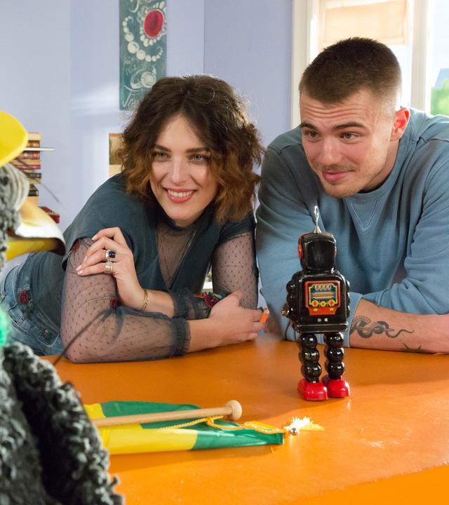 Rudi trägt eine gelb-grüne Kappe und einen gelb-grünen Schal, vor ihm auf dem Tresen legt ein gelb-grüner Wimpel. Er schaut zu Siebenstein und Sommi, vor denen ein Spielzeugroboter steht und die Rudi freundlich anblicken.