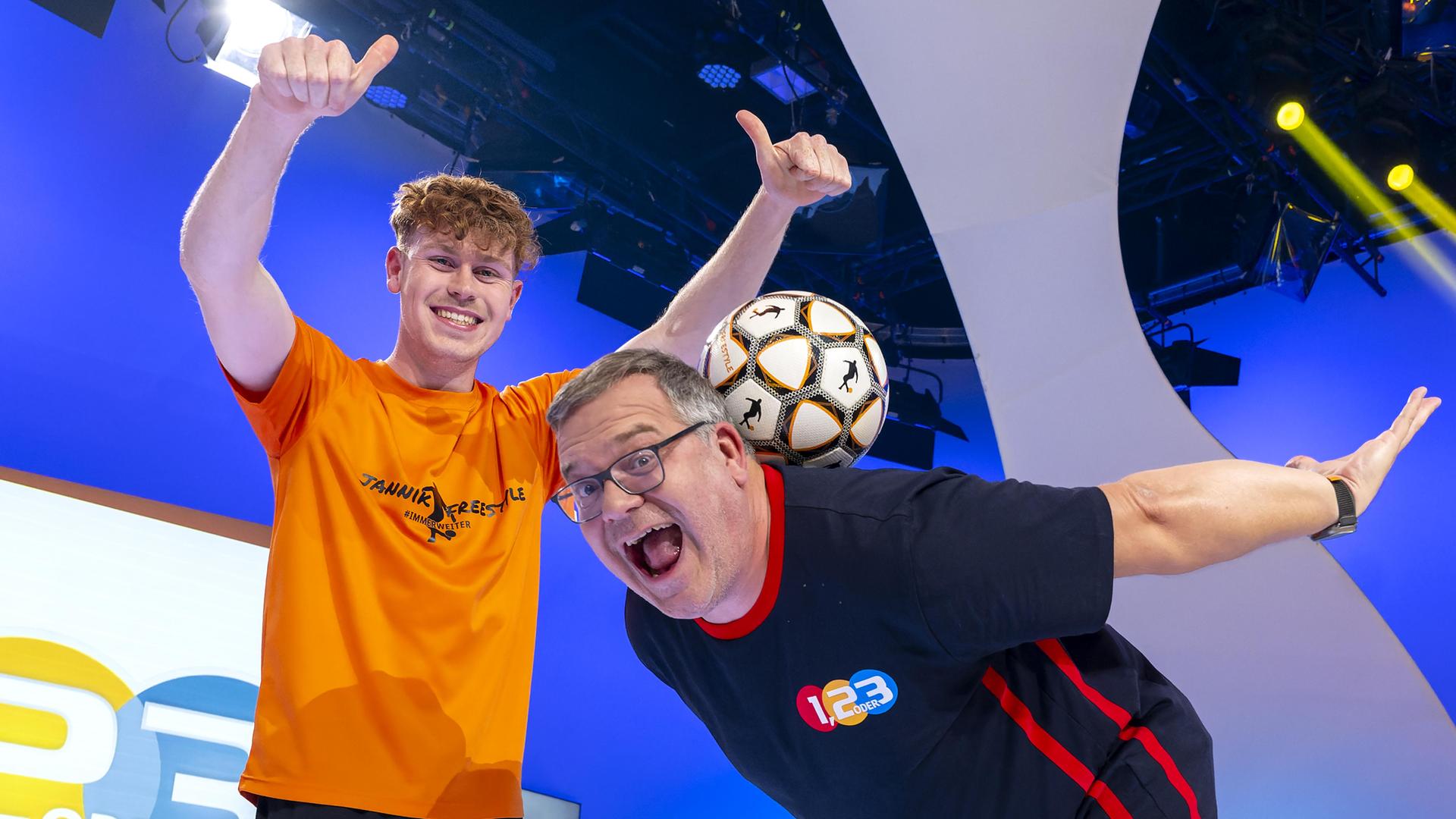 Jannik Freestyle hält beiden Daumen nach oben und grinst, während Elton sich vorn über beugt und versucht einen Fußball im Nacken zu halten.