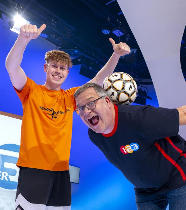 Jannik Freestyle hält beiden Daumen nach oben und grinst, während Elton sich vorn über beugt und versucht einen Fußball im Nacken zu halten.