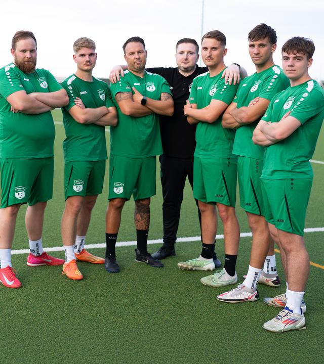 Sechs junge Männer in grünen Fußballtrikots und -hosen stehen mit ihrem Trainer auf einem Fußballfeld.