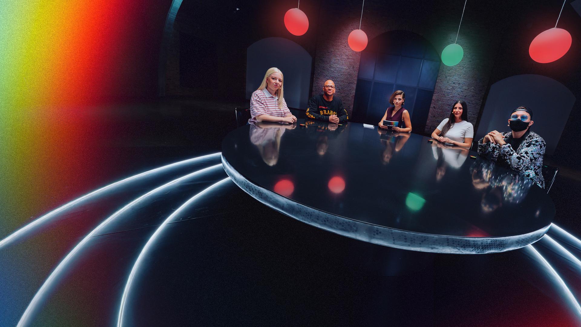 Moderatorin Clara Westhoff (Mitte) sitzt mit den vier Gästen (Jette Nietzard, Etienne Steffen, Meltem Seker und Jay Riddle) der DEEP DEBATE-Folge "Wann geht Cancel Culture zu weit?" an einem großen Runden Tisch. Über ihnen hängen vier runde Lampen, von denen drei rot und eine grün leuchtet.