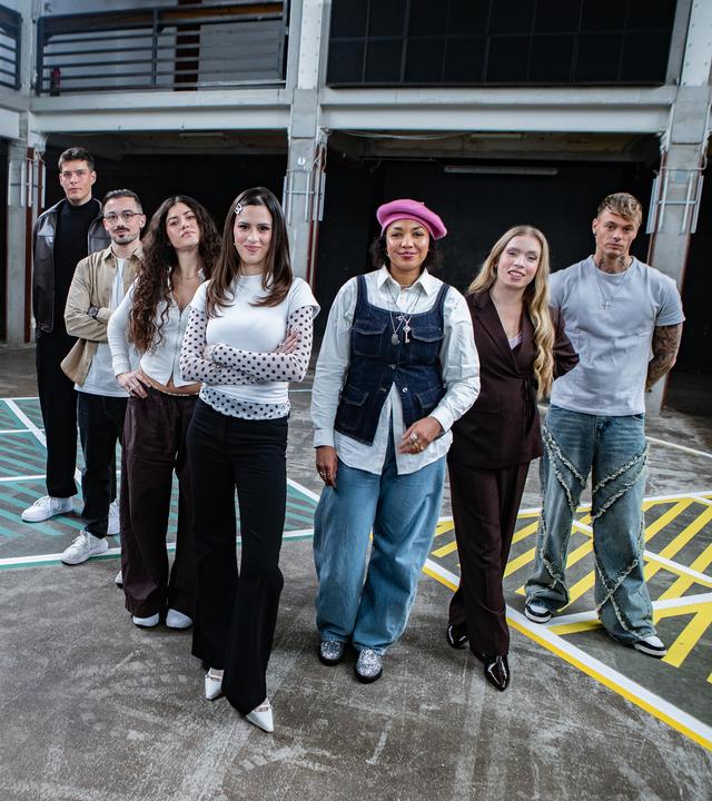 Es stehen sechs Menschen auf einem Boden, der ein schachbrettartiges Muster hat. Die sechs Menschen stehen in einer Reihe. Es sind vier Frauen und drei Männer. Alle blicken in die Richtung des Betrachters.