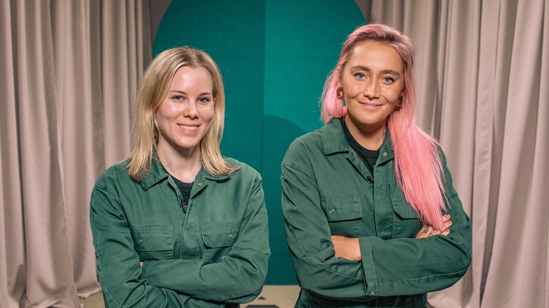 Eine Frau und eine non-binäre Person blicken lächelnd in die Kamera. Sie tragen den gleichen grünen Overall und haben die Arme verschränkt.
