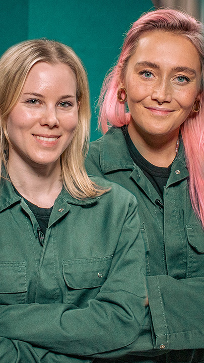 Eine Frau und eine non-binäre Person blicken lächelnd in die Kamera. Sie tragen den gleichen grünen Overall und haben die Arme verschränkt.