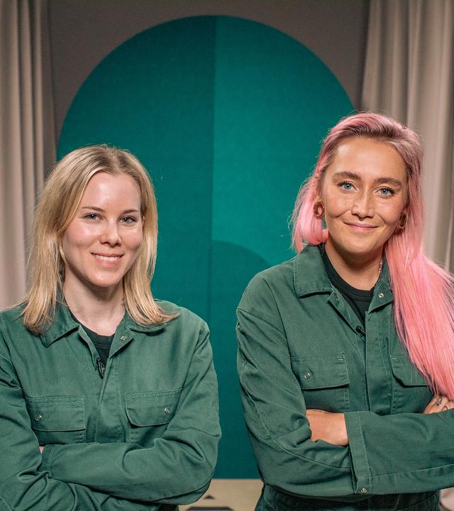 Eine Frau und eine non-binäre Person blicken lächelnd in die Kamera. Sie tragen den gleichen grünen Overall und haben die Arme verschränkt.