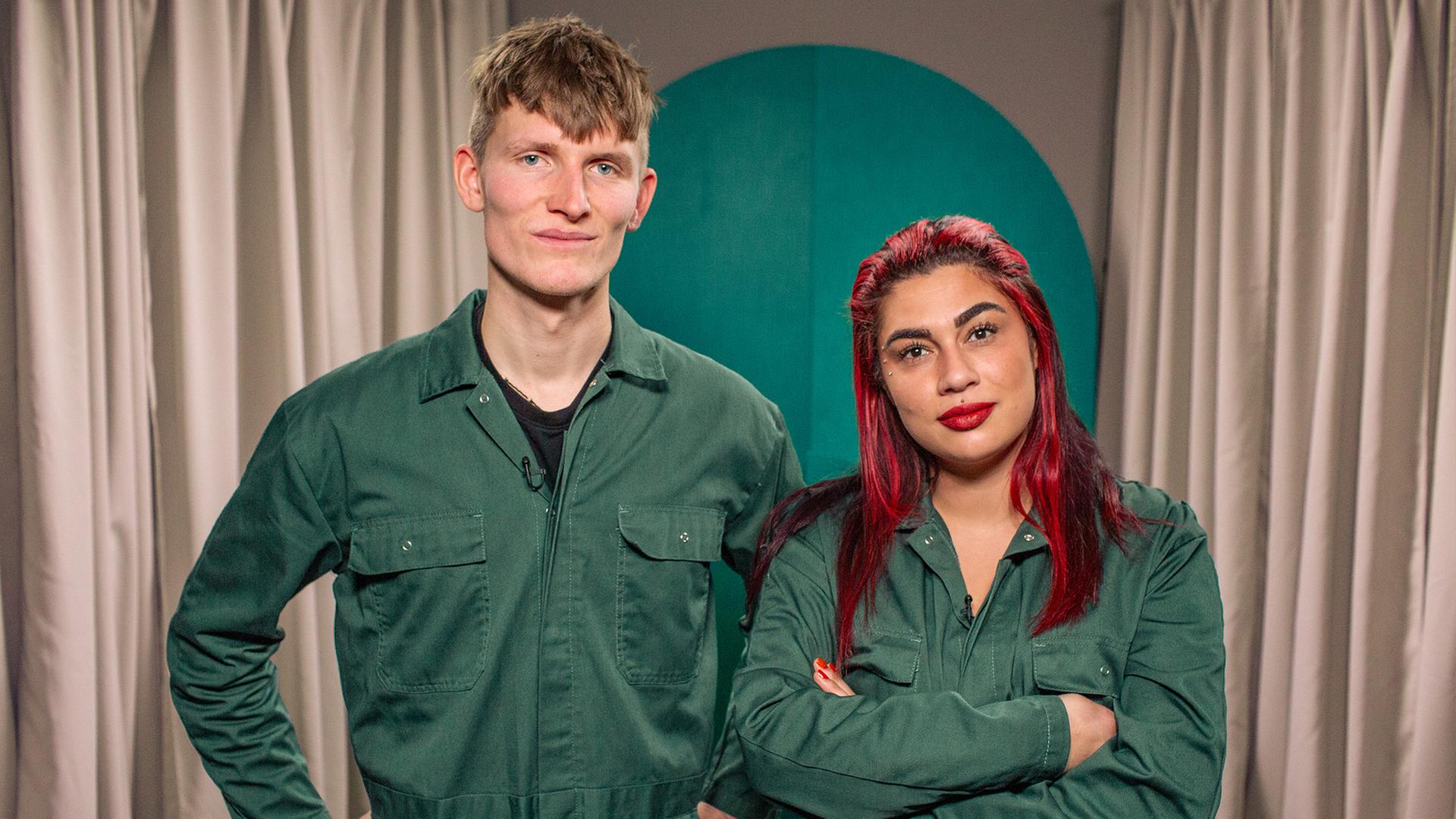 Ein Mann und eine Frau in grünen Overalls stehen nebeneinander und blicken in die Kamera. Der Mann hat die Arme in die Hüfte gestemmt, die Frau hat rote Haare und die Arme verschränkt. Beide lächeln.