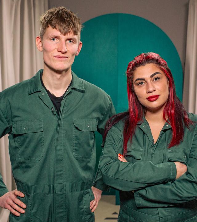 Ein Mann und eine Frau in grünen Overalls stehen nebeneinander und blicken in die Kamera. Der Mann hat die Arme in die Hüfte gestemmt, die Frau hat rote Haare und die Arme verschränkt. Beide lächeln.