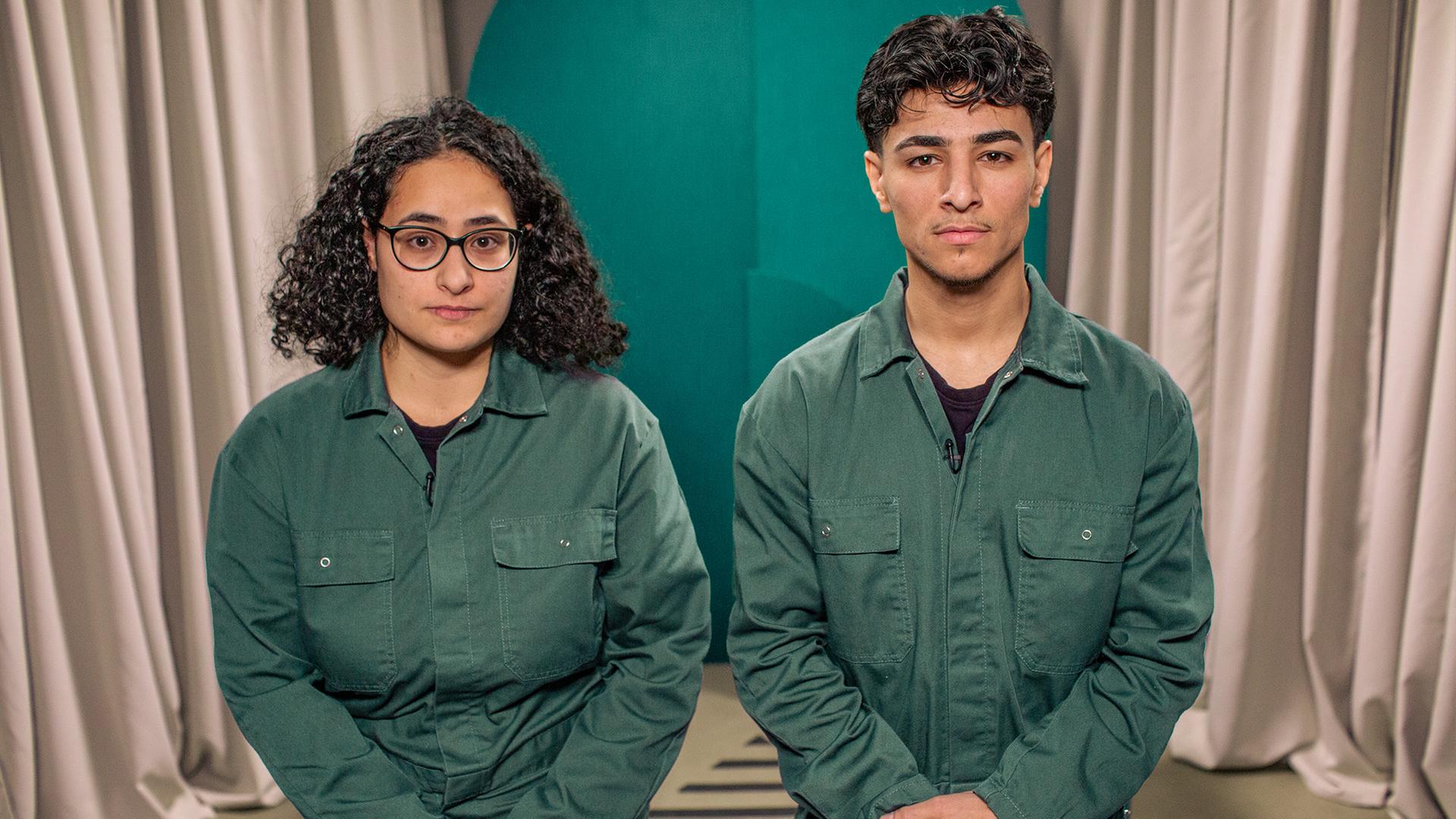 Ein junger Mann und eine junge Frau stehen nebeneinander und blicken in die Kamera. Beide tragen den selben grünen Overall.