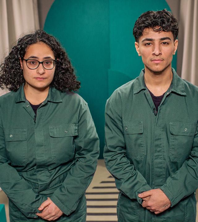 Ein junger Mann und eine junge Frau stehen nebeneinander und blicken in die Kamera. Beide tragen den selben grünen Overall.