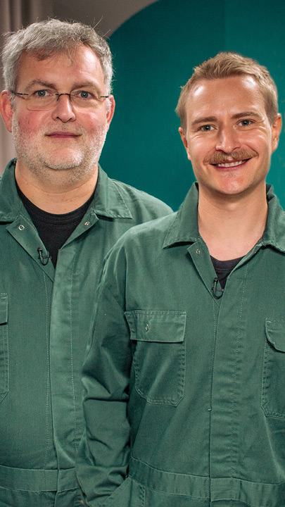 Zwei Männer in grünen Overalls blicken lächelnd in die Kamera.