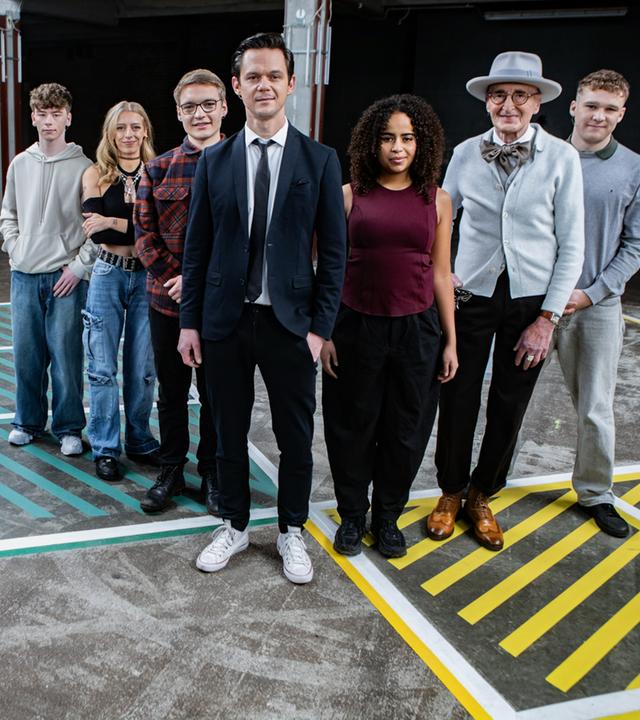 Es stehen sechs Menschen auf einem Boden, der ein schachbrettartiges Muster hat. Die sechs Menschen stehen in einer Reihe. Es sind zwei Frauen und vier Männer. Alle blicken in die Richtung des Betrachters.