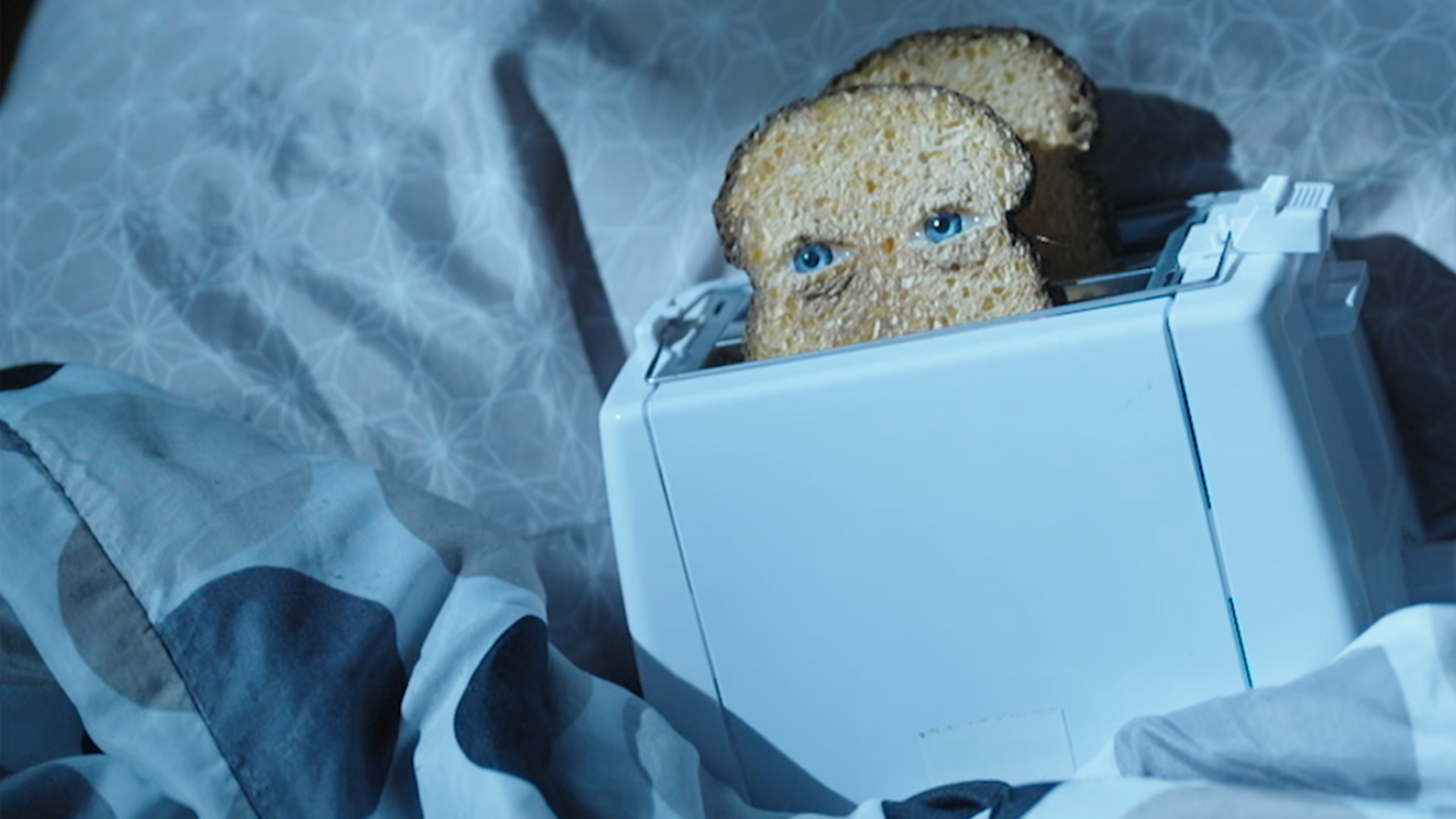 Ein Toaster liegt in einem Bett. Die vordere der beiden Toastscheiben hat Augen.