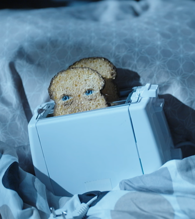 Ein Toaster liegt in einem Bett. Die vordere der beiden Toastscheiben hat Augen.