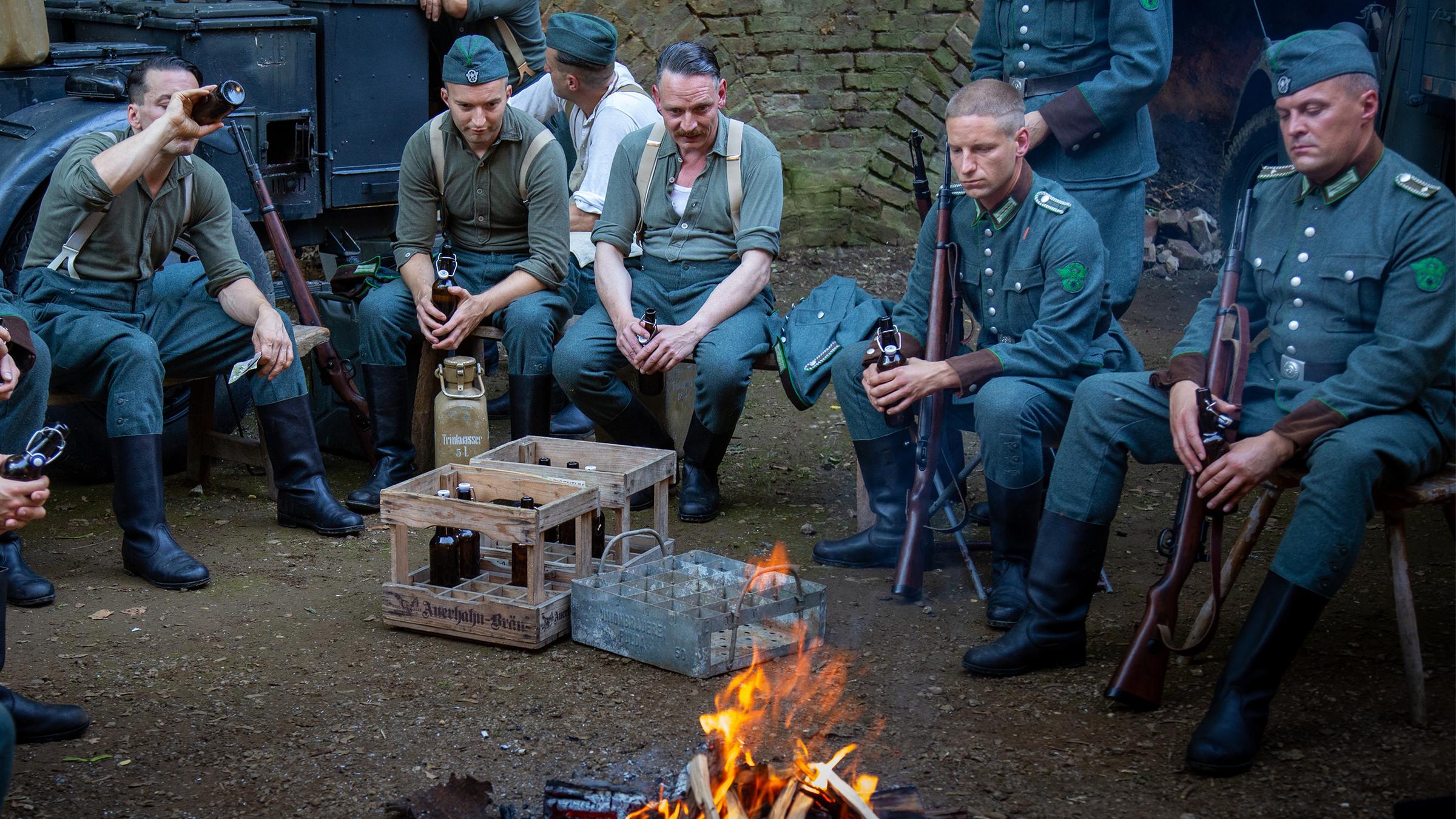 Spielszene: Männer in Uniform, teils mit Gewehren, sitzen am Lagerfeuer.