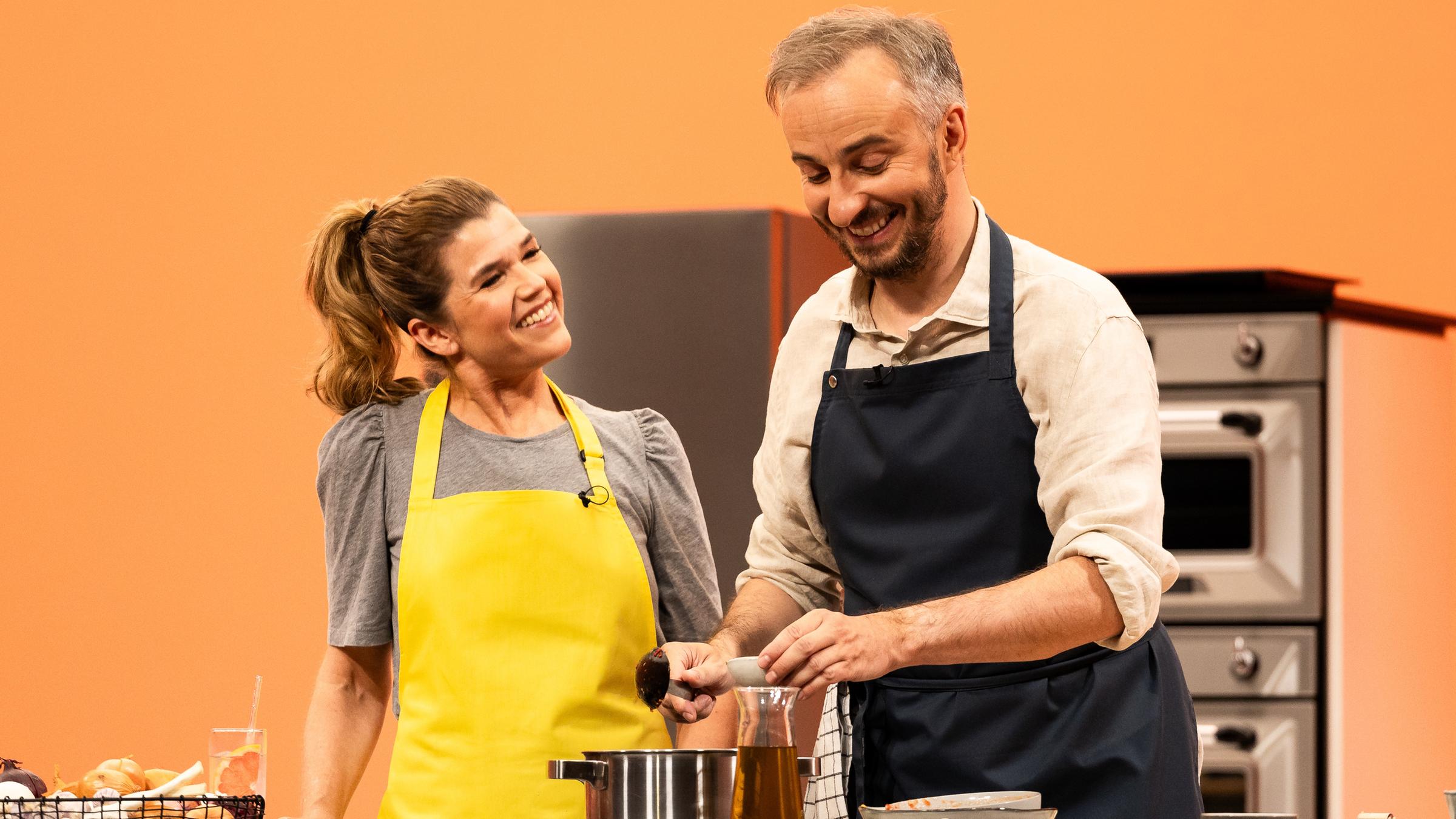 "Böhmi brutzelt": Jan Böhmermann steht mit Anke Engelke in der Küche von "Böhmi brutzelt". Sie kochen gemeinsam. Im Vordergrund steht der Herd. Der Hintergrund ist orange.