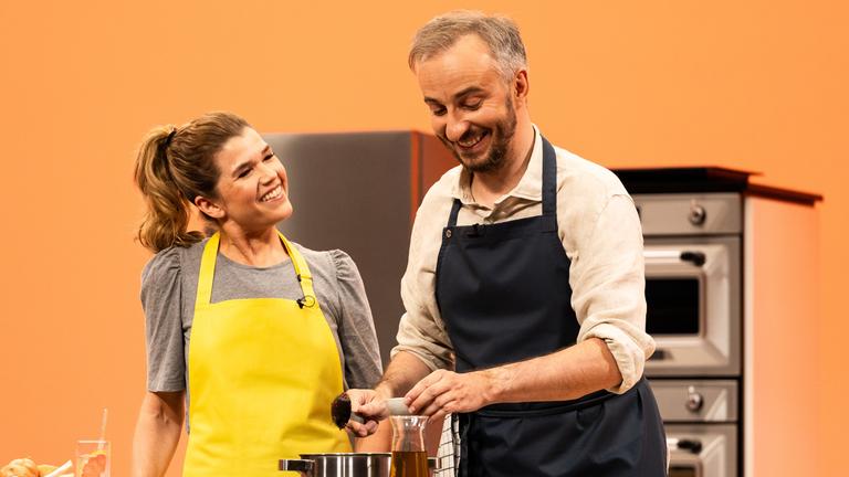 "Böhmi brutzelt": Jan Böhmermann steht mit Anke Engelke in der Küche von "Böhmi brutzelt". Sie kochen gemeinsam. Im Vordergrund steht der Herd. Der Hintergrund ist orange.