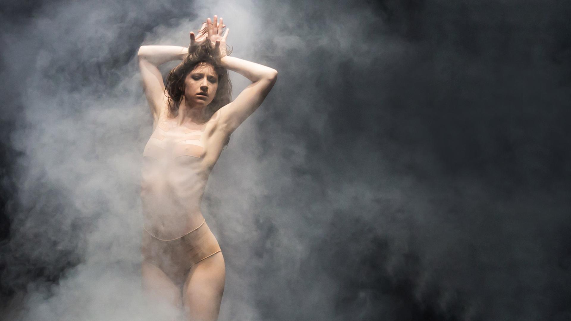 Die Tänzerin steht auf der Bühne, umgeben von Bühnennebel, die Arme verschränkt nach oben gestreckt. Bekleidet ist sie mit einem hautfarbenen Trikot.