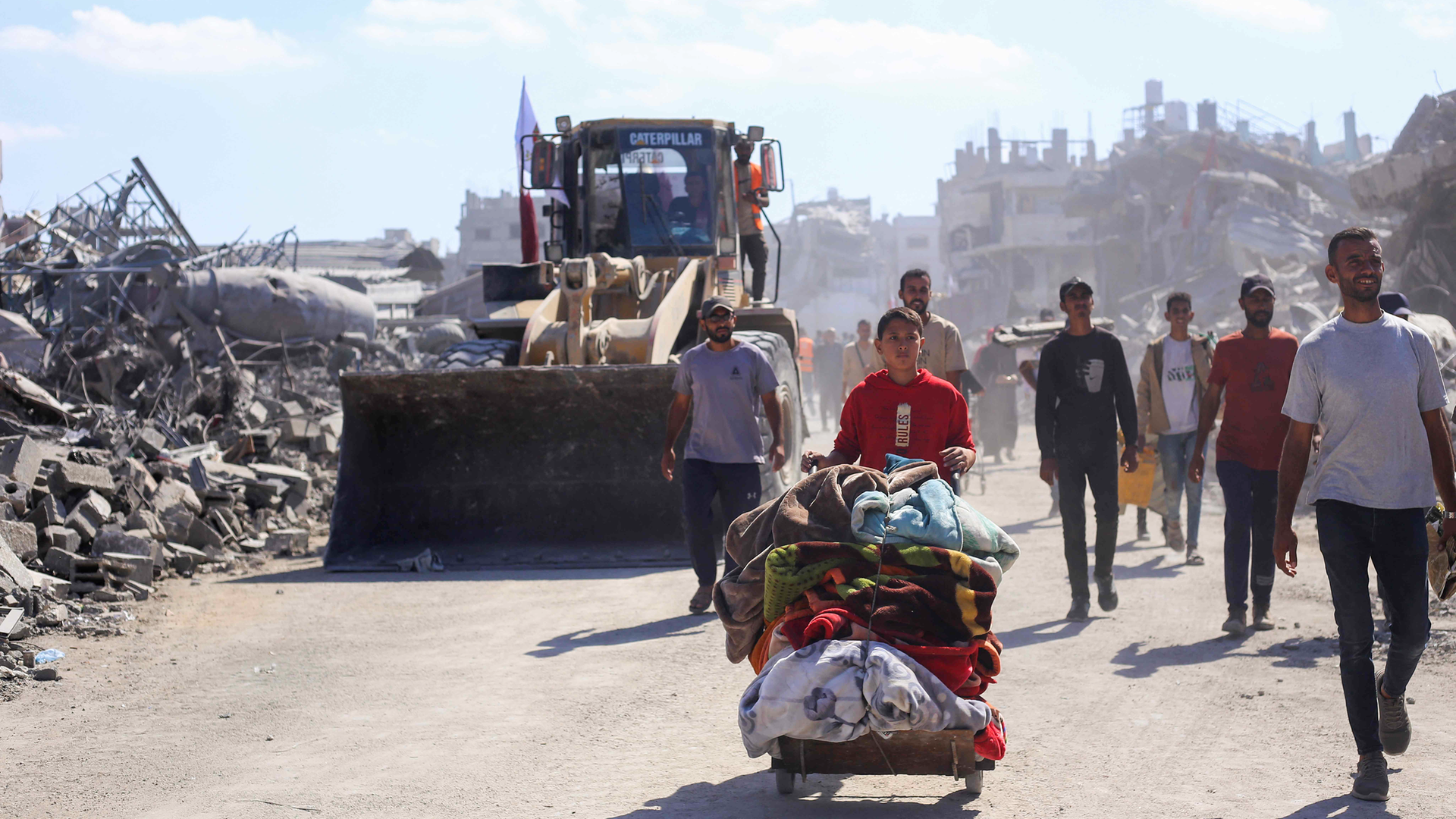 Menschen laufen durch eine zerstörte Stadt, Ein Junge schiebt einen Wagen mit seinen Habseligkeiten, am Straßenrand steht ein Bagger.