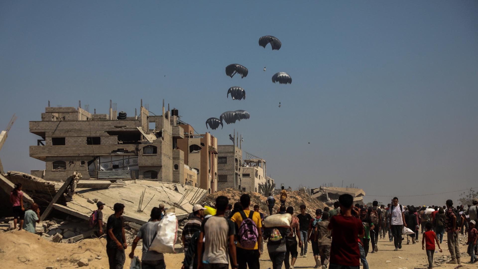 Menschen in Gaza warten am Boden auf herabfallende Fallschirme mit Lebensmittelpaketen. 