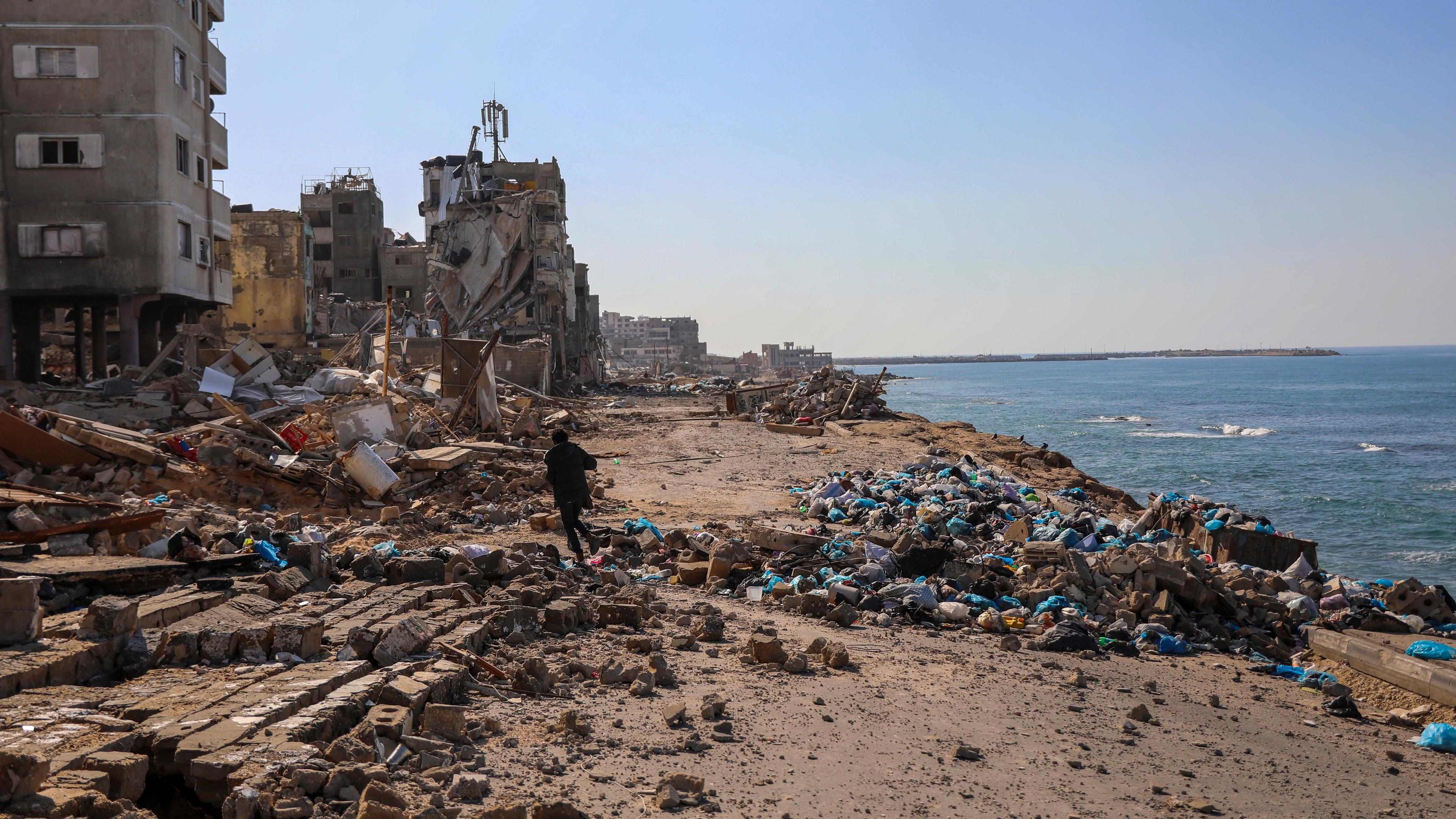 Ein palästinensischer Mann geht am, 29.11.2023, dem sechsten Tag der vorübergehenden Waffenruhe zwischen der Hamas und Israel, am Strand neben zerstörten Gebäuden in Gaza-Stadt spazieren.