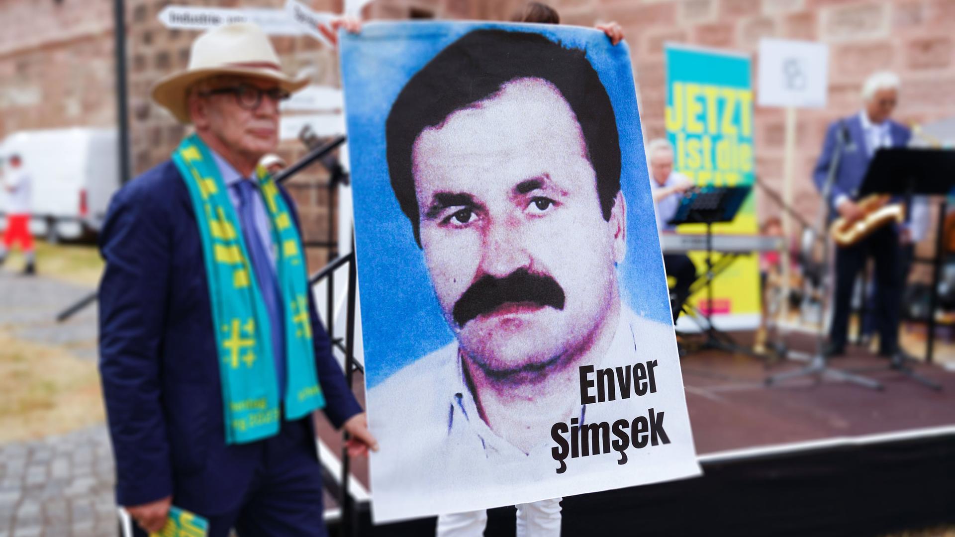 Vor einer Bühne hält eine Person ein großes Plakat mit dem Portätfoto eines Mannes mit Schnauzbart hoch.