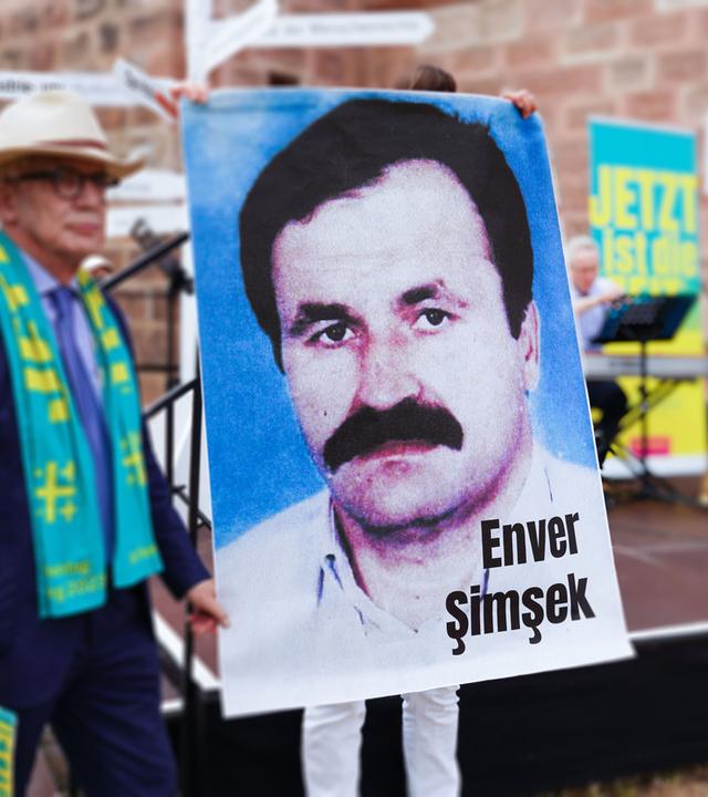 Vor einer Bühne hält eine Person ein großes Plakat mit dem Portätfoto eines Mannes mit Schnauzbart hoch.