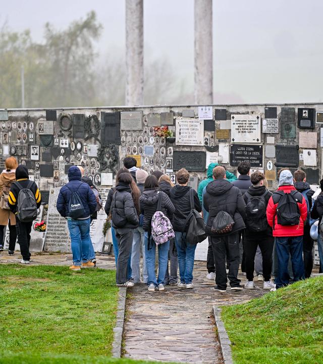Schüler stehen im Freien vor einer Mauer, an der viele Tafeln angebracht sind.