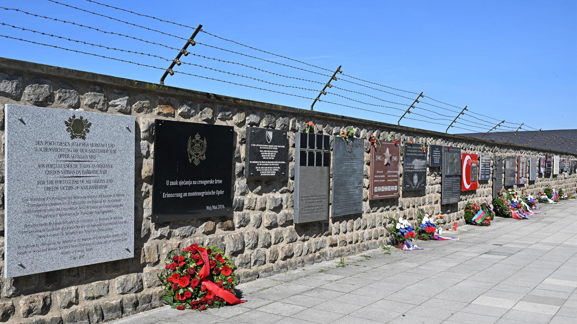 Eine Mauer mit einem Stacheldraht darüber. An der ganzen Mauer entlang hängen Gedenktafeln in verschiedenen Farben und Größen, alle sind rechteckig. Vor einigen Tafeln auf dem Boden liegen Blumenkränze.