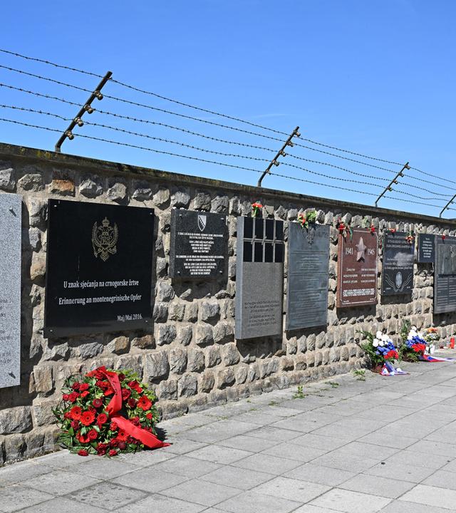 Eine Mauer mit einem Stacheldraht darüber. An der ganzen Mauer entlang hängen Gedenktafeln in verschiedenen Farben und Größen, alle sind rechteckig. Vor einigen Tafeln auf dem Boden liegen Blumenkränze.