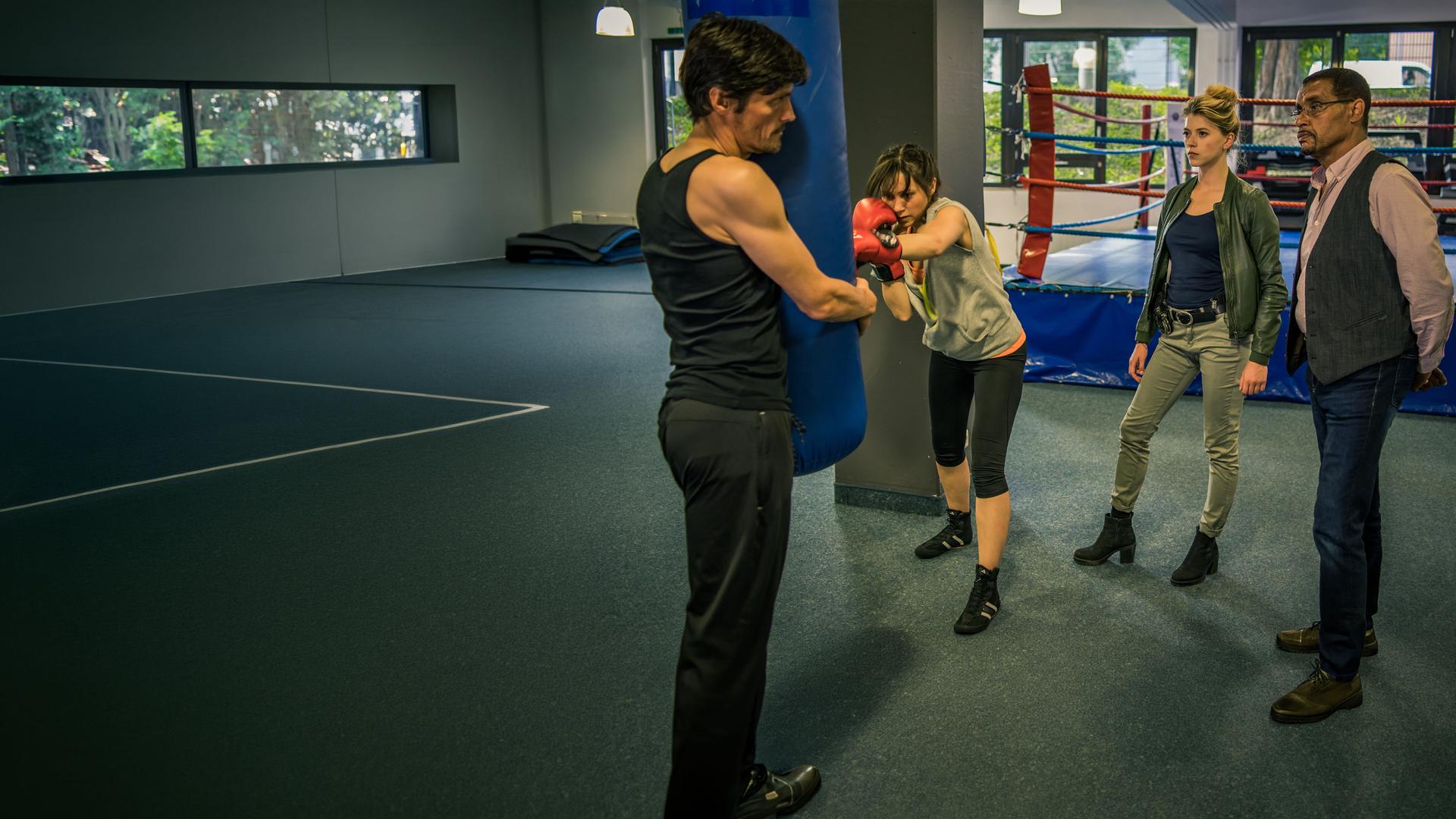 "SOKO München - Gefährlicher Dunstkreis": Franz Ainfachnur (Christofer v. Beau) und Resi Schwaiger (Mersiha Husagic) stehen in einer Sporthalle vor einem Boxring und beobachten mit ernstem Blick Stanislaw Koslowski (Stipe Erceg) und Tilda Wager (Celia Hoffmann), die vor ihnen an einem Boxsack trainieren. Tilda trägt Boxhandschuhe und schlägt auf den Sack. Stanislaw Koslowski hält den Sack von der anderen Seite aus fest und blickt angespannt zu den beiden Kommissaren.