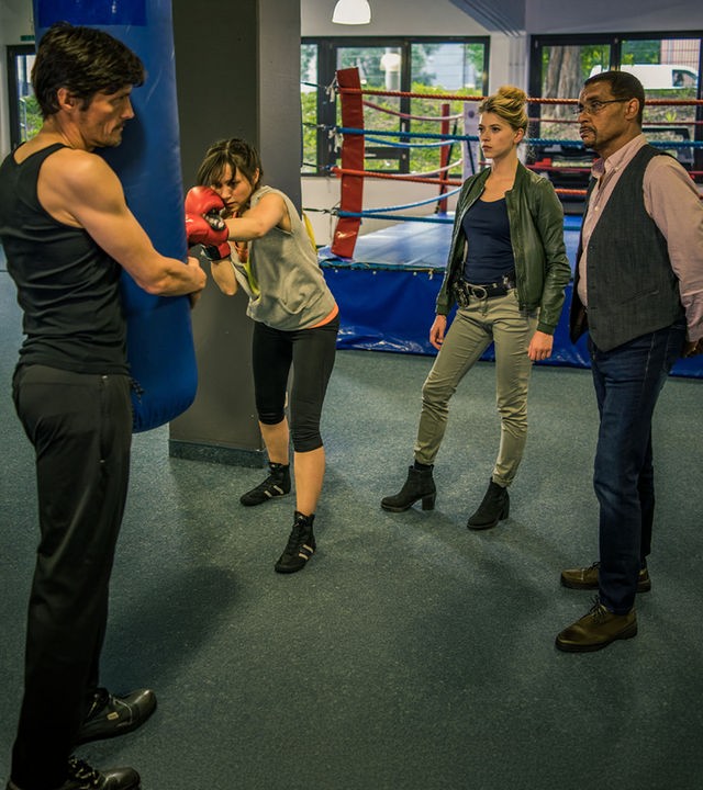 "SOKO München - Gefährlicher Dunstkreis": Franz Ainfachnur (Christofer v. Beau) und Resi Schwaiger (Mersiha Husagic) stehen in einer Sporthalle vor einem Boxring und beobachten mit ernstem Blick Stanislaw Koslowski (Stipe Erceg) und Tilda Wager (Celia Hoffmann), die vor ihnen an einem Boxsack trainieren. Tilda trägt Boxhandschuhe und schlägt auf den Sack. Stanislaw Koslowski hält den Sack von der anderen Seite aus fest und blickt angespannt zu den beiden Kommissaren.