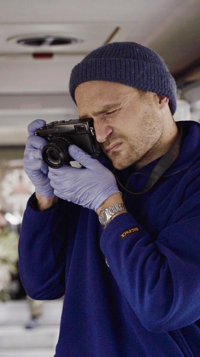 In einem Bus, der mit Liegen und medizinischem Equipment speziell für Krankentransporte ausgestattet wurde, steht Vincent Haiges und fotografiert, ein Auge zugekniffen, das andere durch den Sucher seiner Kamera schauend. Er trägt einen dicken, blauen Pullover, Wollmütze und medizinische Handschuhe. Neben ihm hängt ein Infusionsbeutel der einen Soldaten versorgt. Weiter hinten liegen weitere verwundete Soldaten.