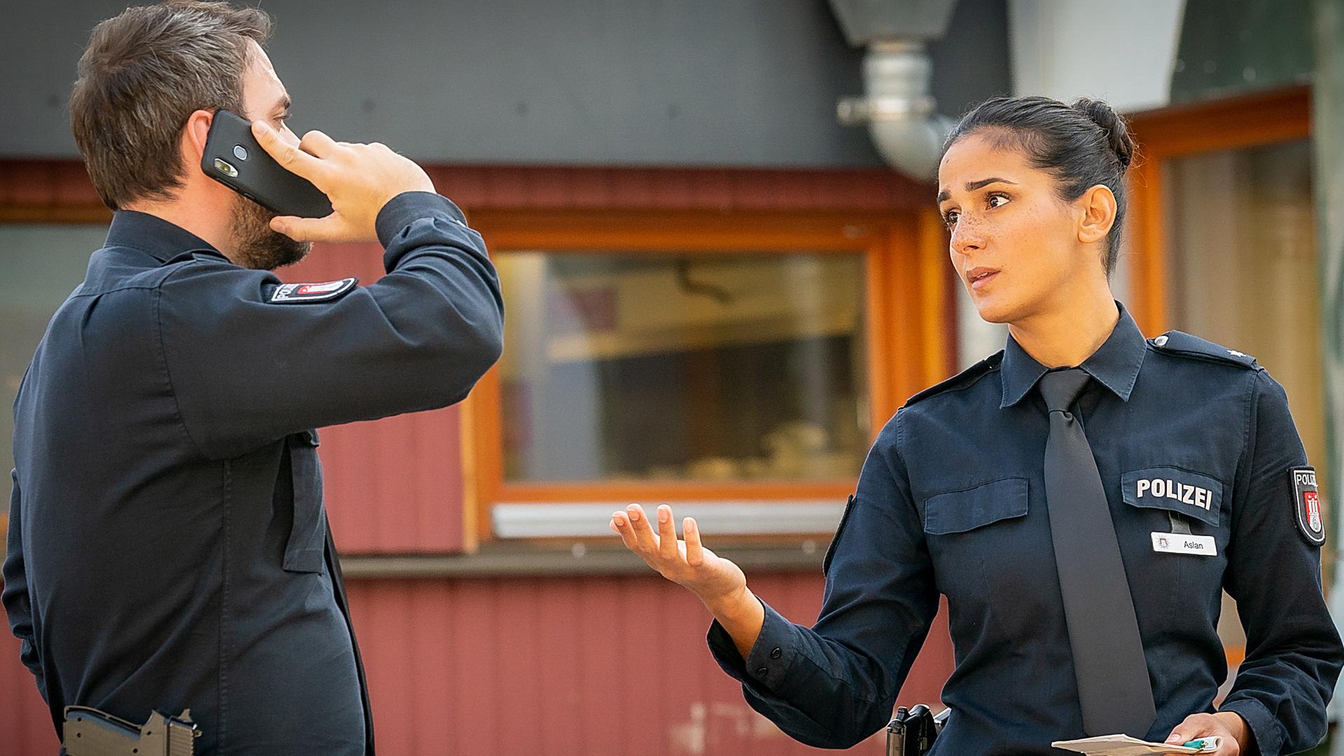  Der Polizist Kris Freiberg (Marc Barthel) steht vor einem Gebäude und telefoniert. Neben ihm steht seine Kollegin Pinar Aslan (Aybi Era). Sie sieht ihn an. In ihrer linken Hand hält sie einen Notizblock und einen Stift. Mit der Rechten gestikuliert sie.