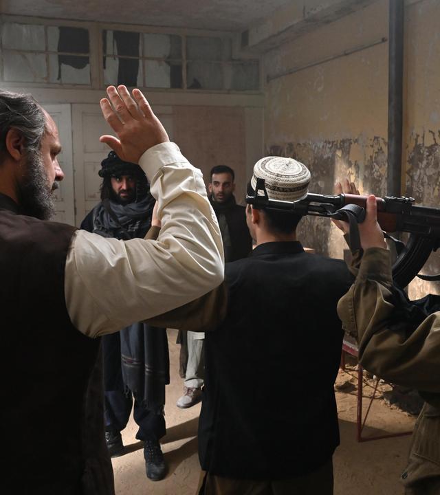 In einem heruntergekommenen Raum heben zwei Männer die Hände hoch, die von Taliban mit Waffen bedroht werden.