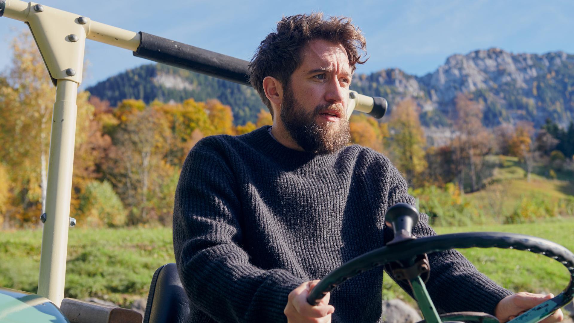 Nahaufnahme von Schorschi Striebel (Sebastian Edtbauer), der mit einem Traktor fährt. Sein Gesichtsausdruck ist verzweifelt. Im Hintergrund Bergpanorama.