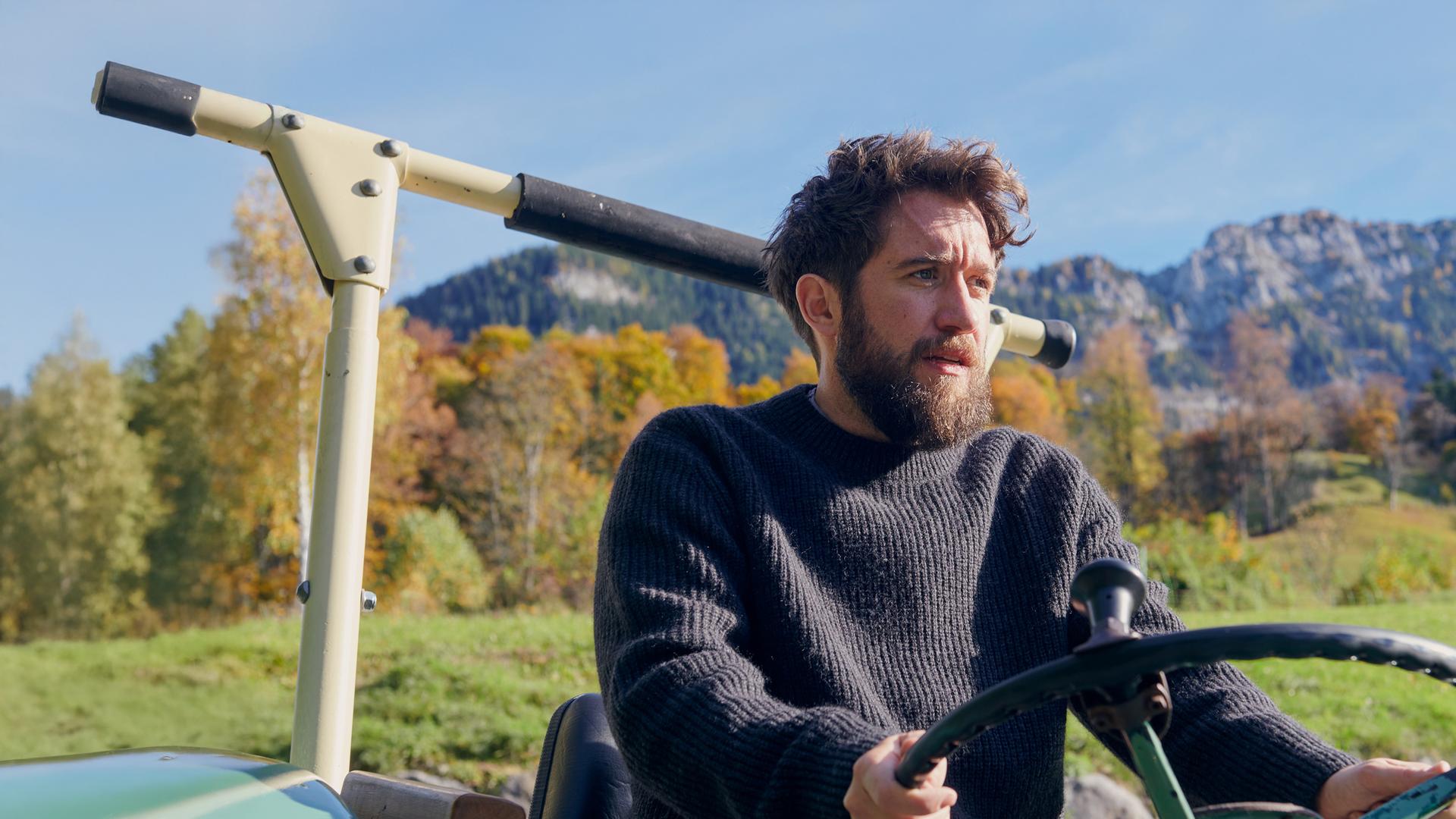 Nahaufnahme von Schorschi Striebel (Sebastian Edtbauer), der mit einem Traktor fährt. Sein Gesichtsausdruck ist verzweifelt. Im Hintergrund Bergpanorama.
