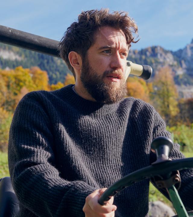 Nahaufnahme von Schorschi Striebel (Sebastian Edtbauer), der mit einem Traktor fährt. Sein Gesichtsausdruck ist verzweifelt. Im Hintergrund Bergpanorama.