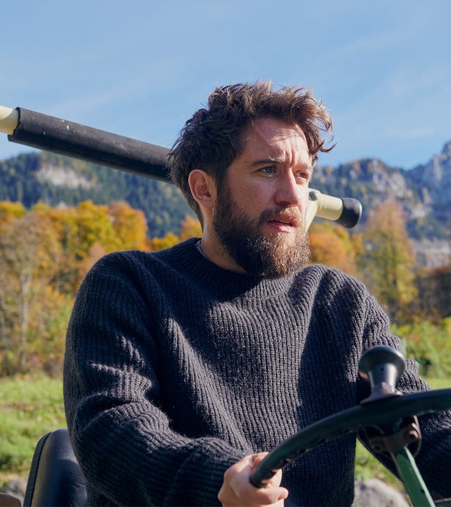 Nahaufnahme von Schorschi Striebel (Sebastian Edtbauer), der mit einem Traktor fährt. Sein Gesichtsausdruck ist verzweifelt. Im Hintergrund Bergpanorama.
