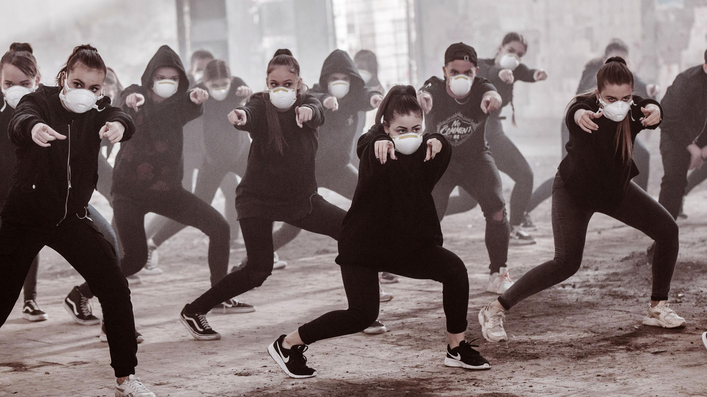 Eine Gruppe schwarz gekleideter Tänzer*innen mit weißen FFP2-Masken bei Tanz