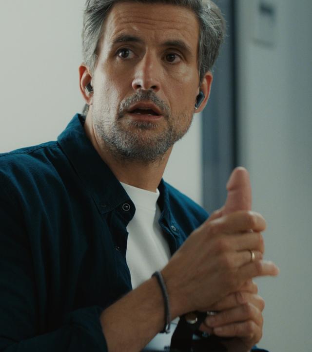 "The other gAIrl – … gegessen wird zuhause": Frank (Tom Beck) sitzt in seinem Büro und hält einen Dildo in der Hand.