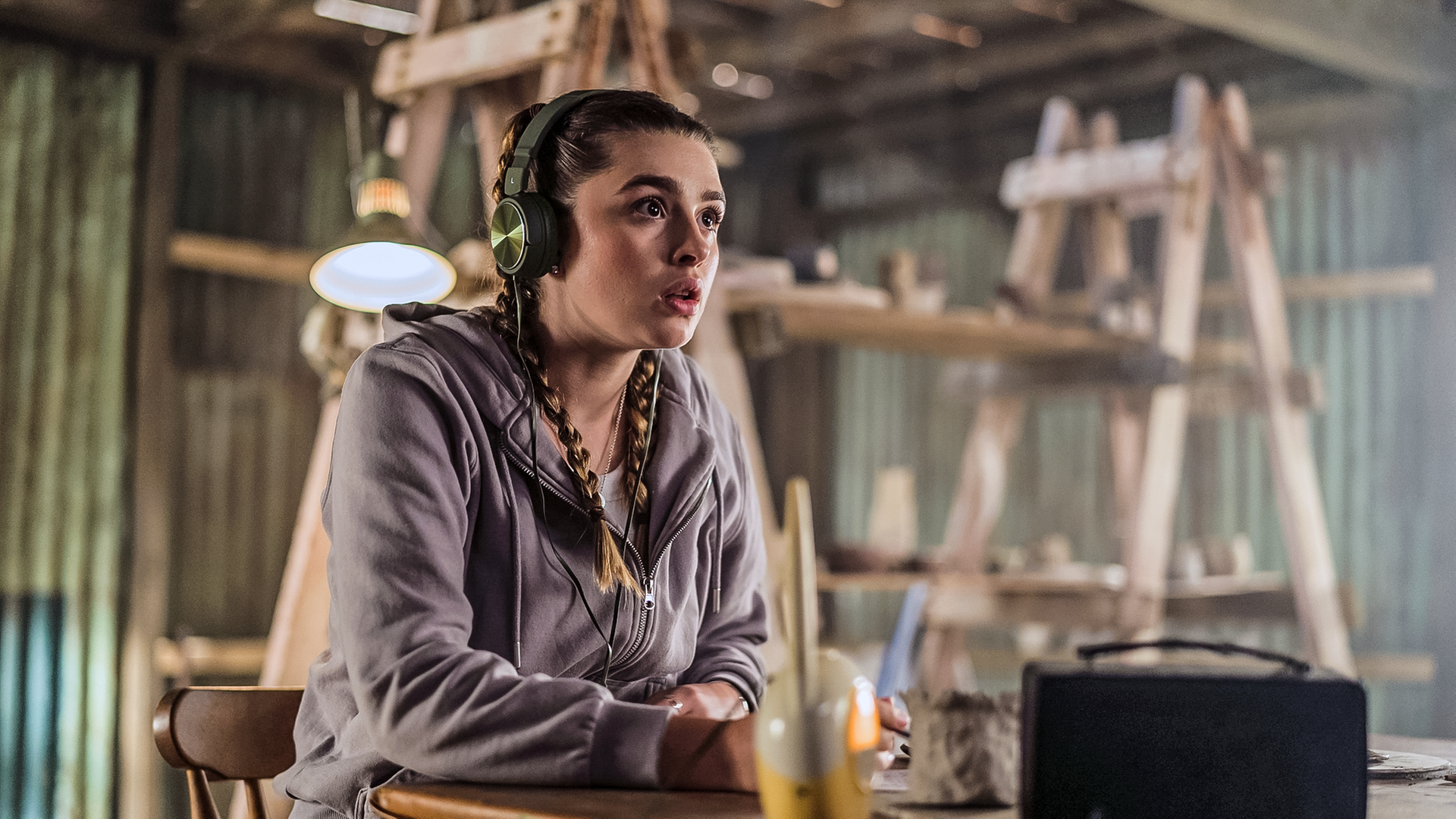 Natasha sitzt konzentriert zuhörend mit Kopfhörern auf den Ohren und einem Babyphone an einem Tisch und belauscht ein Gespräch.