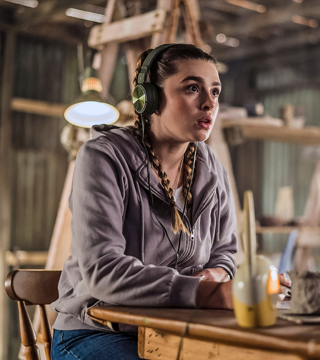 Natasha sitzt konzentriert zuhörend mit Kopfhörern auf den Ohren und einem Babyphone an einem Tisch und belauscht ein Gespräch.